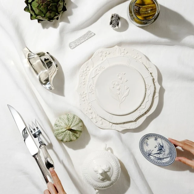 Overhead shot of white dinnerware, artichoke, silver cutlery, and a woman's hand holding a decorative plate on a white cloth background.
