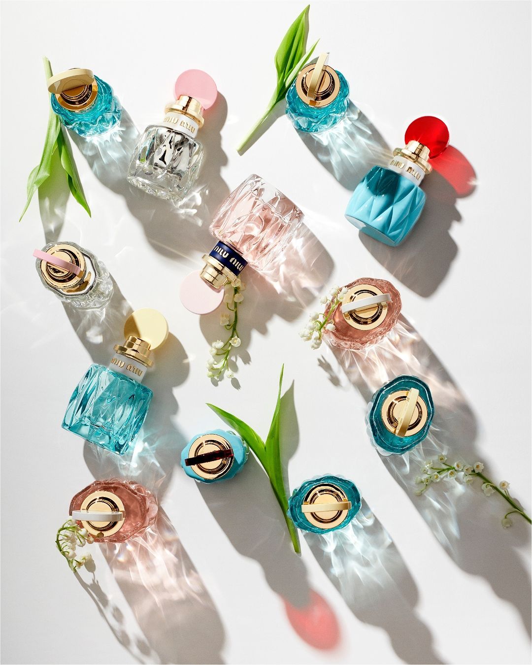 Overhead shot showcases colorful Miu Miu perfume bottles on a white surface, accented by scattered green leaves and delicate white flowers.
