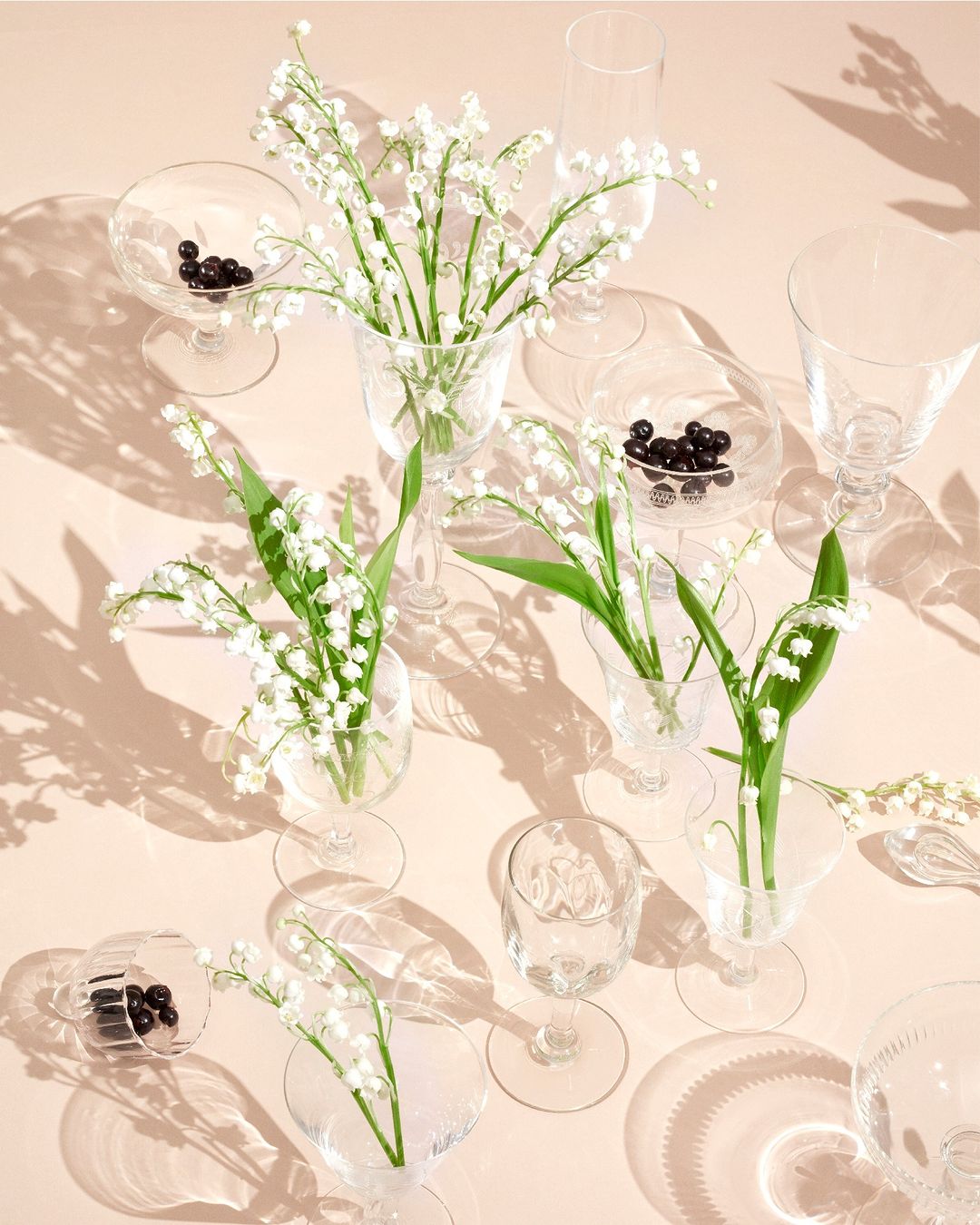 Overhead shot of lily of the valley flowers in clear glasses, accented by chocolate spheres, casting shadows on a peach surface.