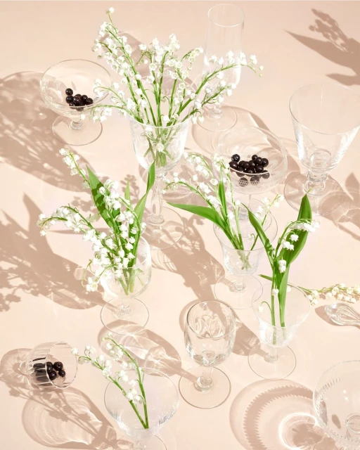 Overhead shot of lily of the valley flowers in clear glasses, accented by chocolate spheres, casting shadows on a peach surface.