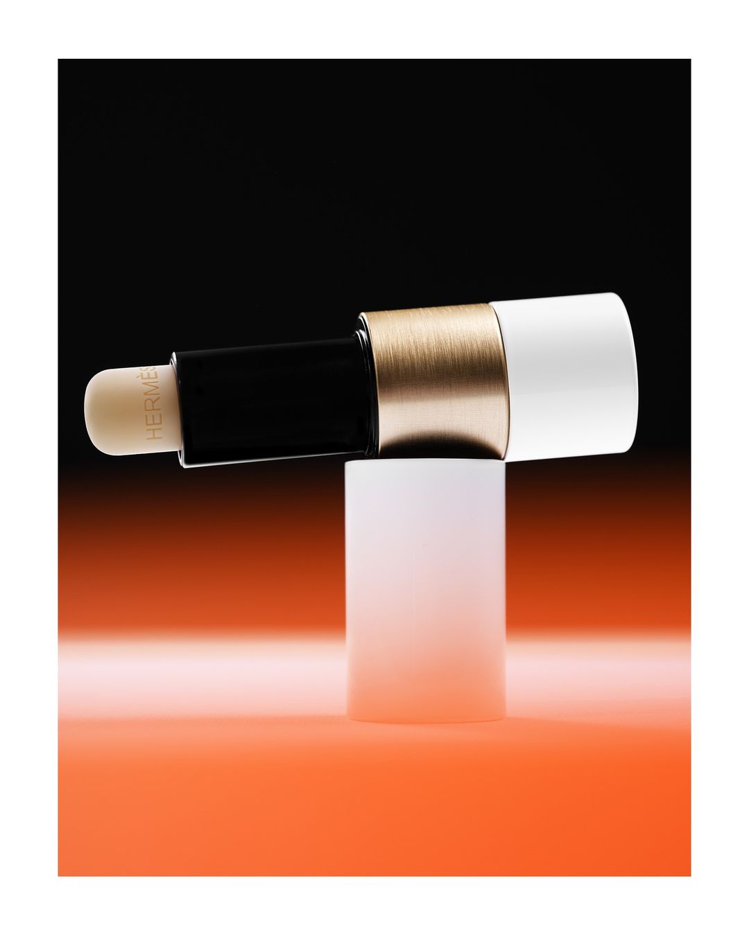 Hermes lipstick tube in black, gold, and white on a white pedestal against a split orange and black background in studio lighting.