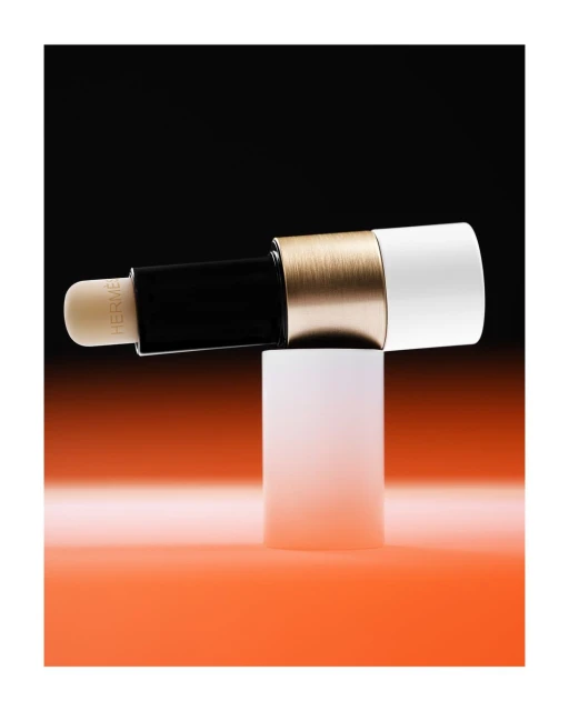 Hermes lipstick tube in black, gold, and white on a white pedestal against a split orange and black background in studio lighting.