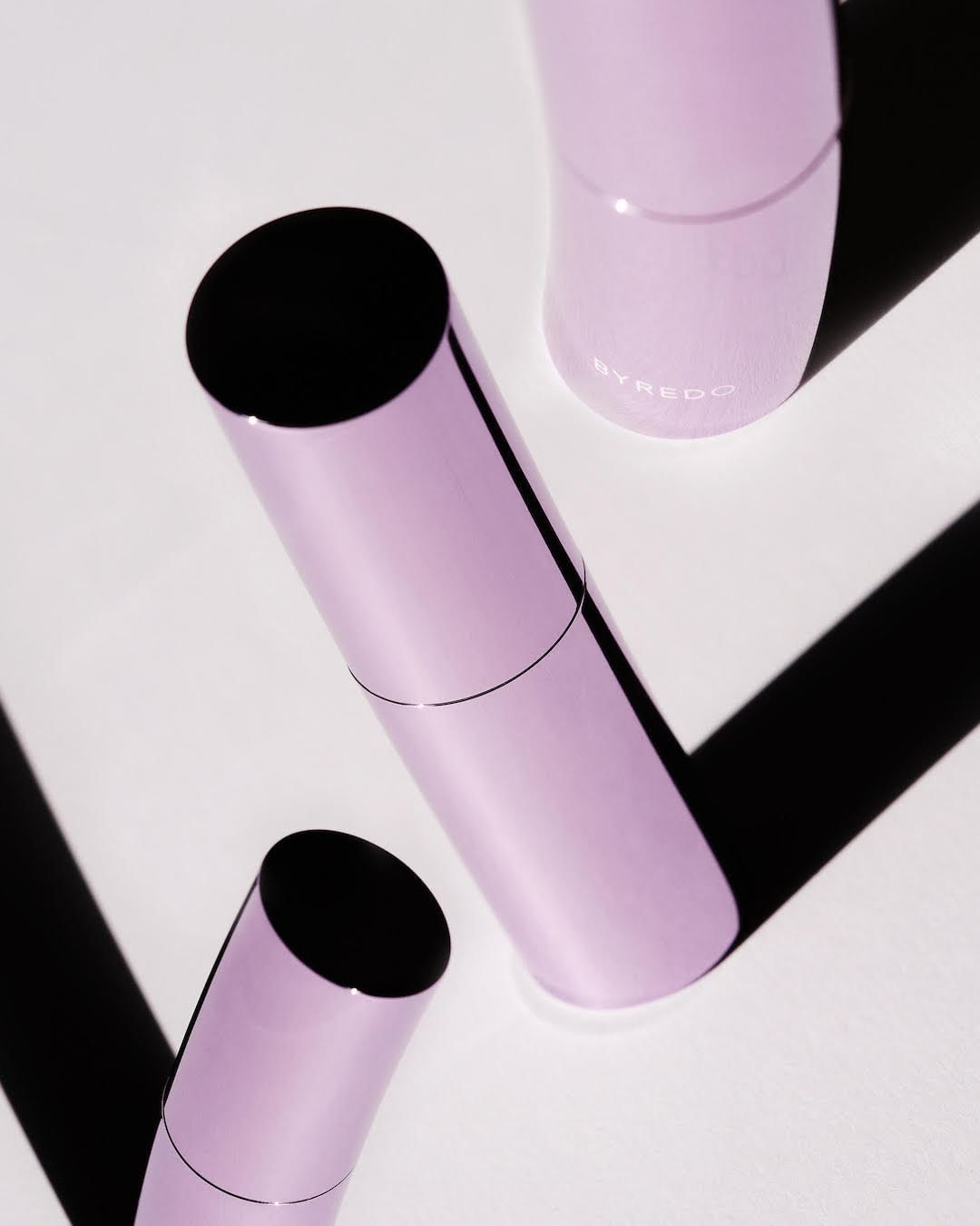 Three cylindrical lilac-colored perfume bottles with black caps, arranged diagonally on a white surface with strong shadows.