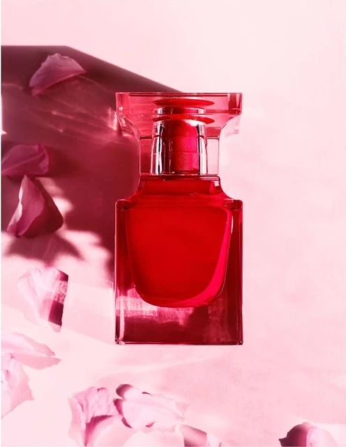 Crystal perfume bottle with red liquid, surrounded by scattered pink flower petals on a light pink background and soft shadows.