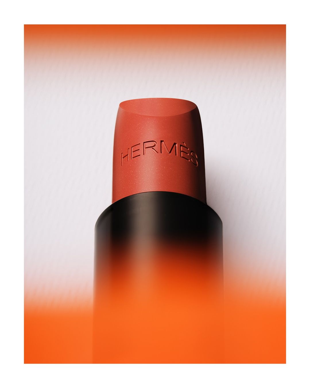 Close-up of an orange Hermès lipstick tube with the brand name embossed, against a blurred orange and white gradient background.
