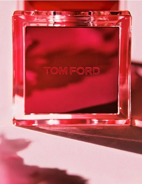 Close-up of a reflective, square Tom Ford fragrance bottle with red accents and clear edges, casting soft shadows on a light surface.