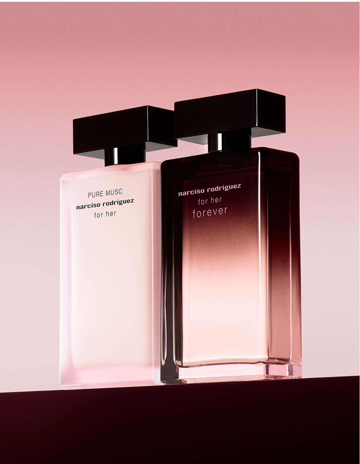 Two Narciso Rodriguez perfume bottles, 'Pure Musc' and 'For Her Forever,' stand on a dark surface against a soft pink backdrop.