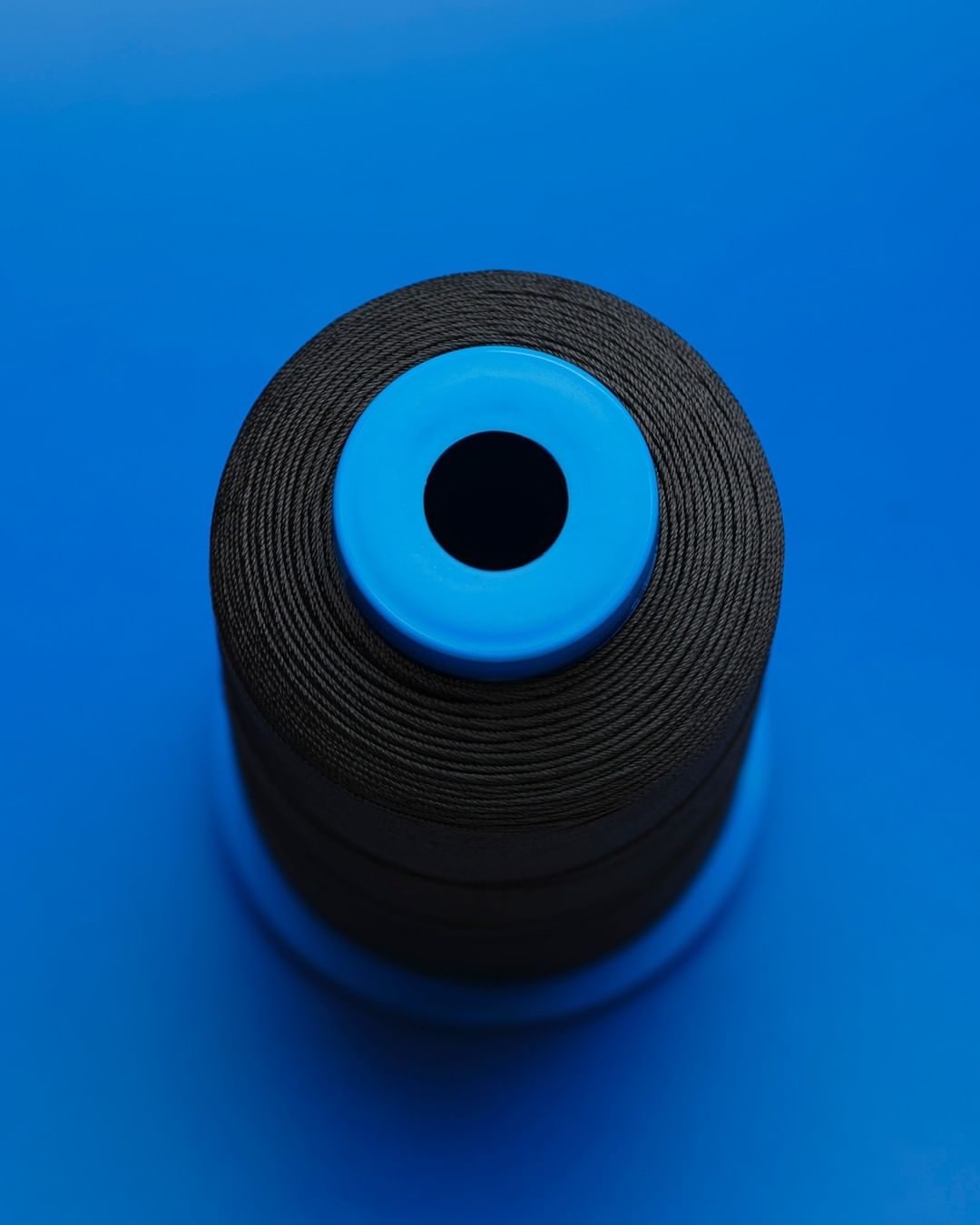 Overhead studio shot of a spool of black thread with a blue plastic core, set against a vibrant blue background. Close-up, detailed texture.