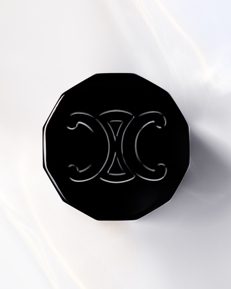 Elevated view of an octagonal black cosmetic compact with a silver decorative logo on a bright white surface with light refraction.