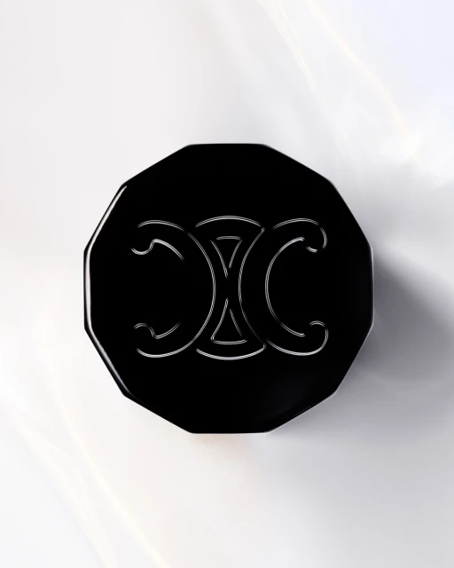Elevated view of an octagonal black cosmetic compact with a silver decorative logo on a bright white surface with light refraction.