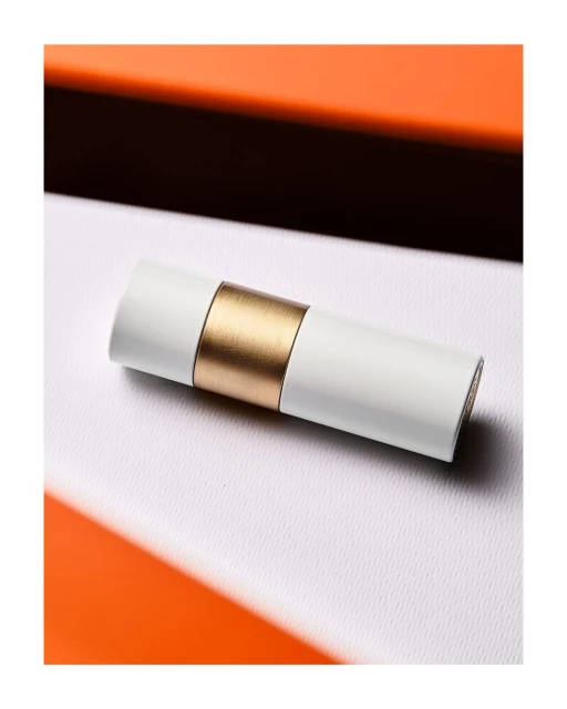 Close-up shot of a cylindrical white cosmetic container with a gold band on a white surface with an orange background element.