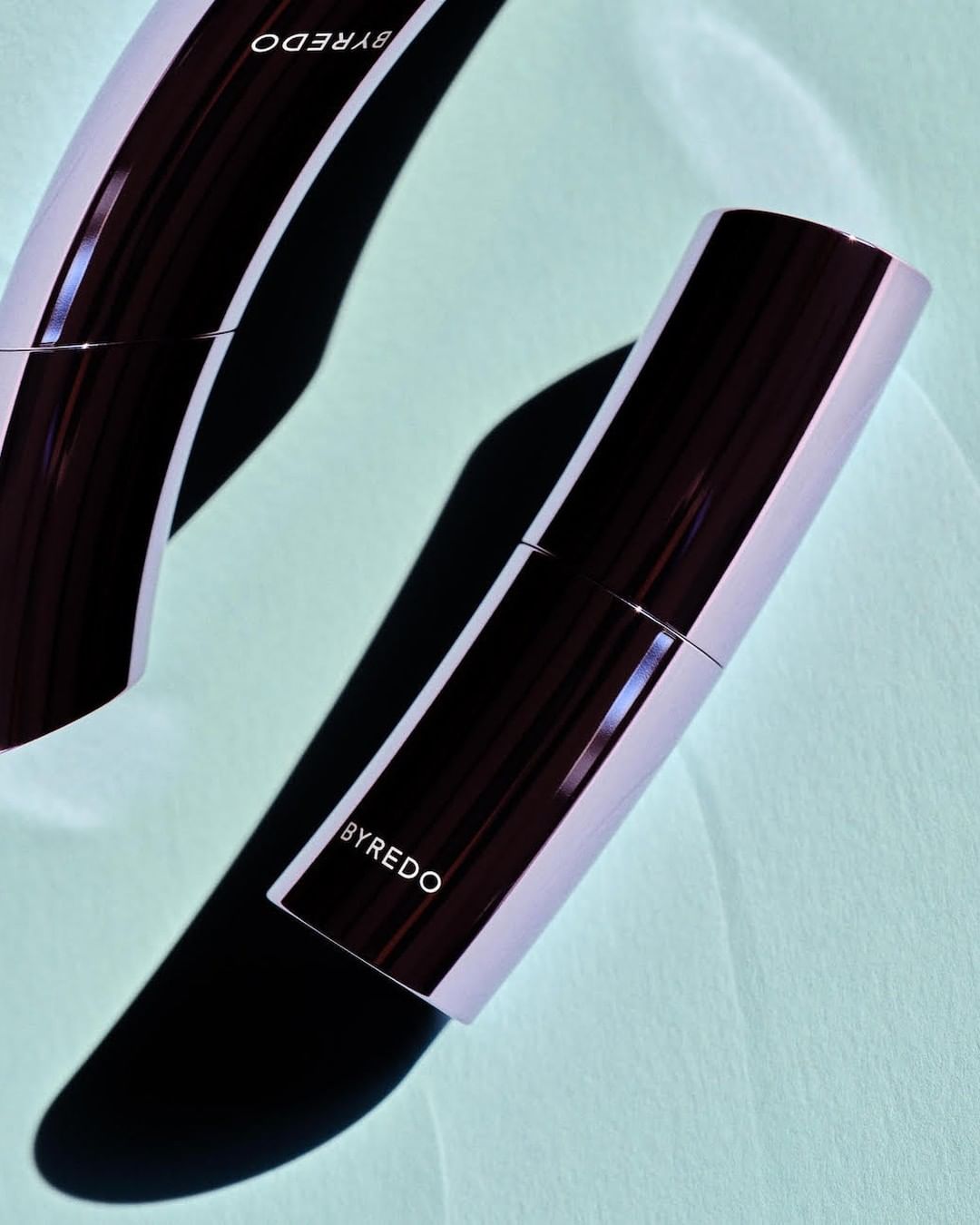 Close-up product shot of two Byredo cosmetic sticks on a light teal surface with soft shadows, emphasizing beauty and minimalist design.
