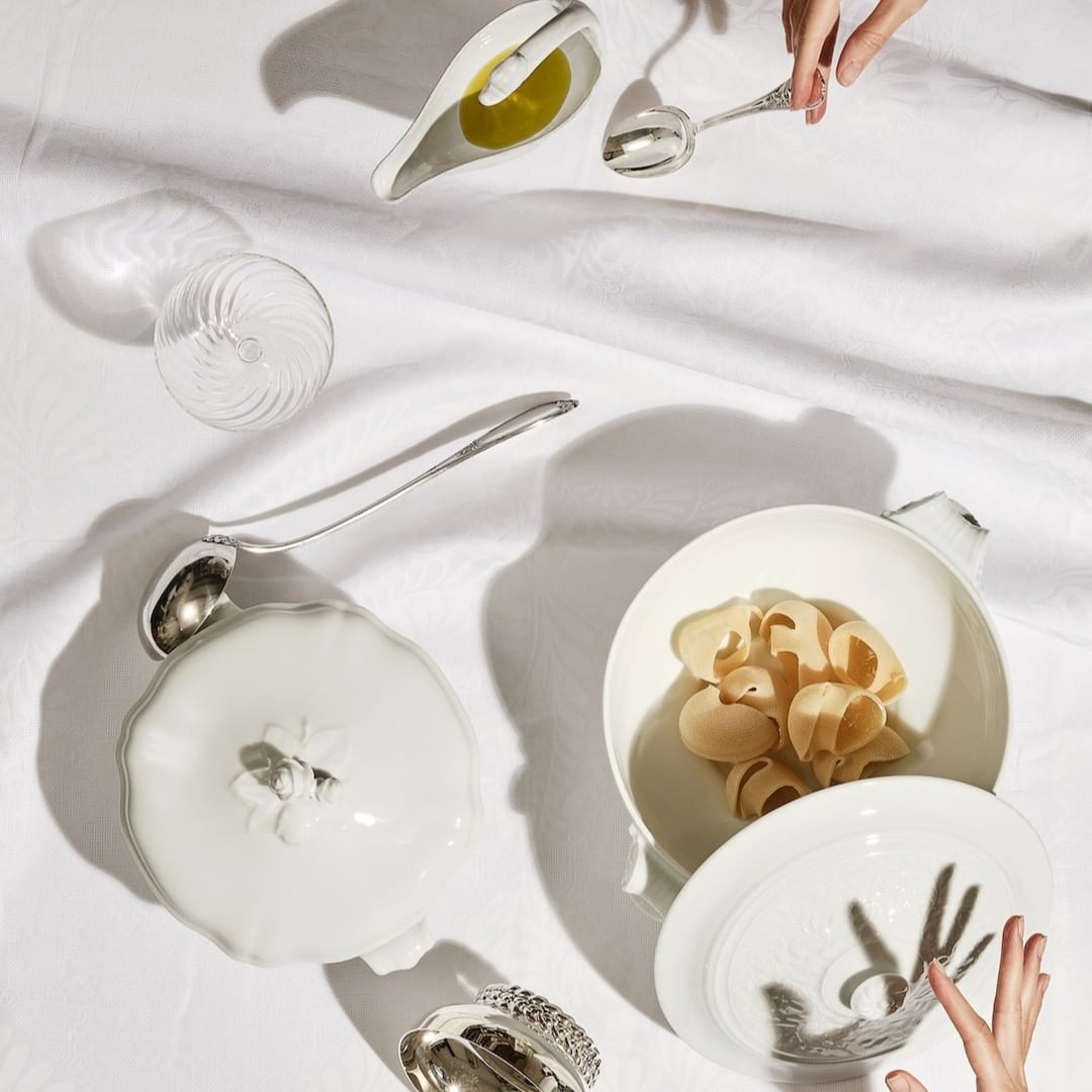 Overhead shot of pasta shells in white ceramic bowl, silver spoons, glass, and oil on white linen, lit with dramatic shadows.