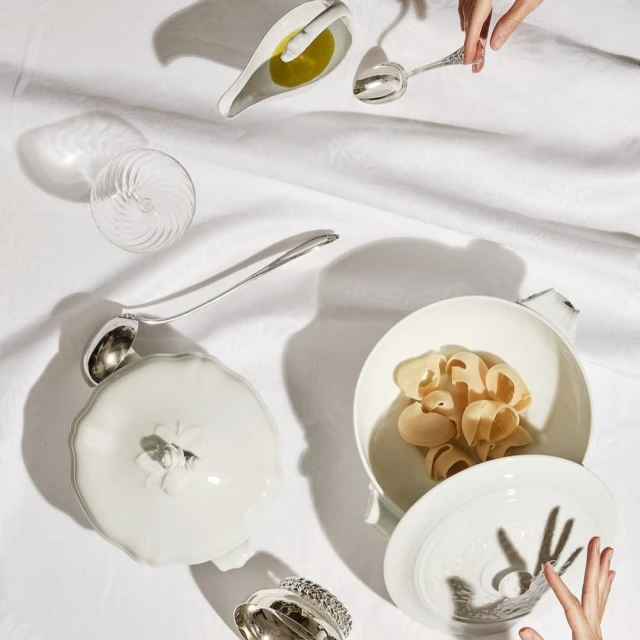 Overhead shot of pasta shells in white ceramic bowl, silver spoons, glass, and oil on white linen, lit with dramatic shadows.