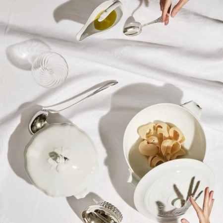 Overhead shot of pasta shells in white ceramic bowl, silver spoons, glass, and oil on white linen, lit with dramatic shadows.