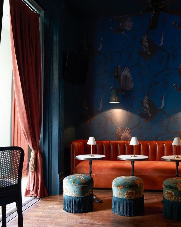 Interior of a moody room with a coral couch and teal ottomans, set against dark blue crane-patterned wallpaper, softly lit.