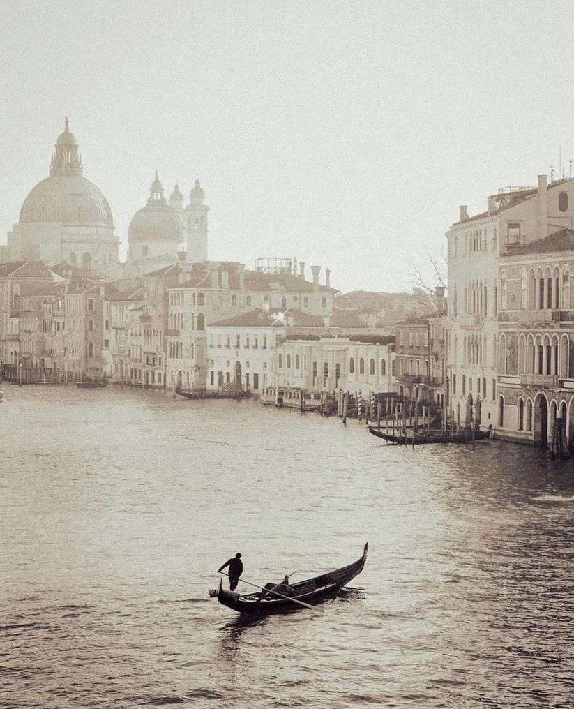 Sepia image of a gondola with two people navigating a canal in Venice, Italy, with buildings and a domed church in the background.