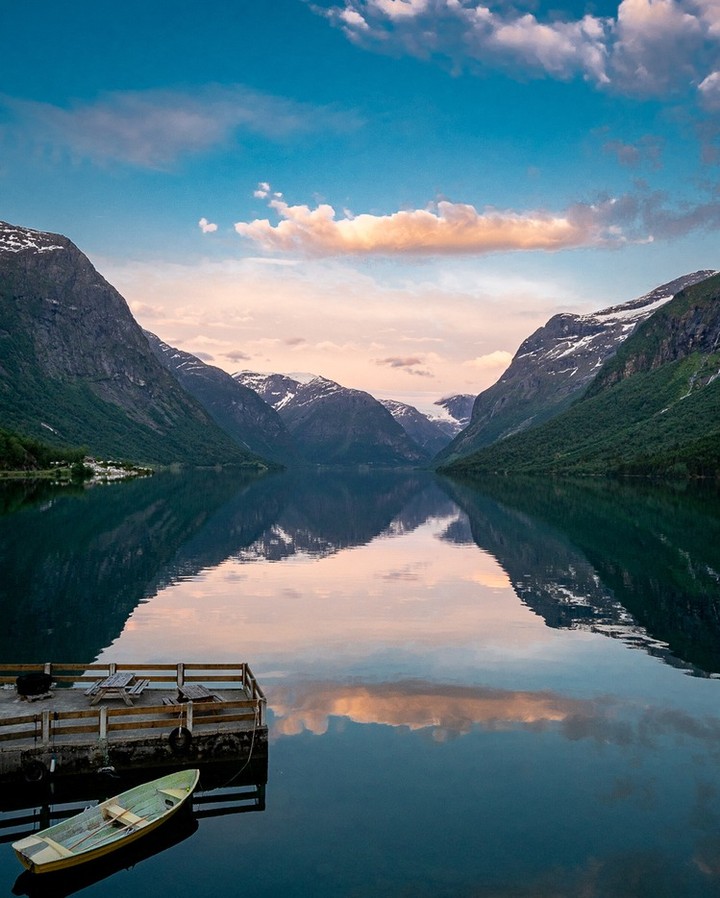 Scenic view of a calm lake reflecting mountains and sky, with a wooden dock and small boat in the foreground at dusk or dawn.