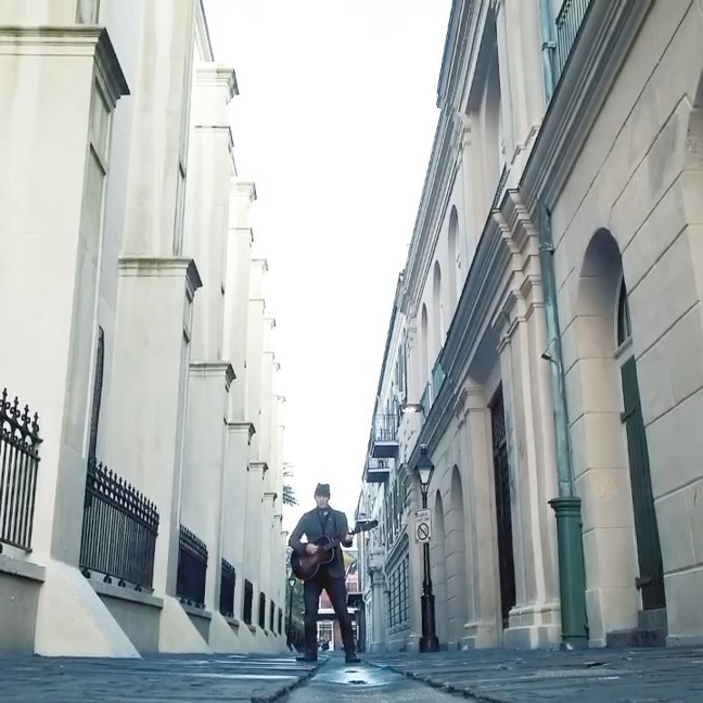 Adult male playing acoustic guitar on cobblestone street in city. Building exteriors and street lamp visible on either side.
