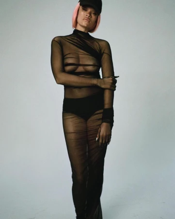 Model in sheer black dress and pink wig wearing a black cap against a plain light background