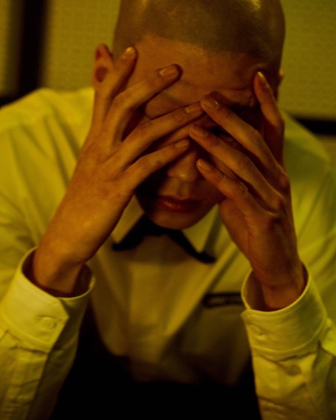 Close-up of a bald adult male in a white dress shirt and black bow tie with his face obscured by his hands in a photo studio.