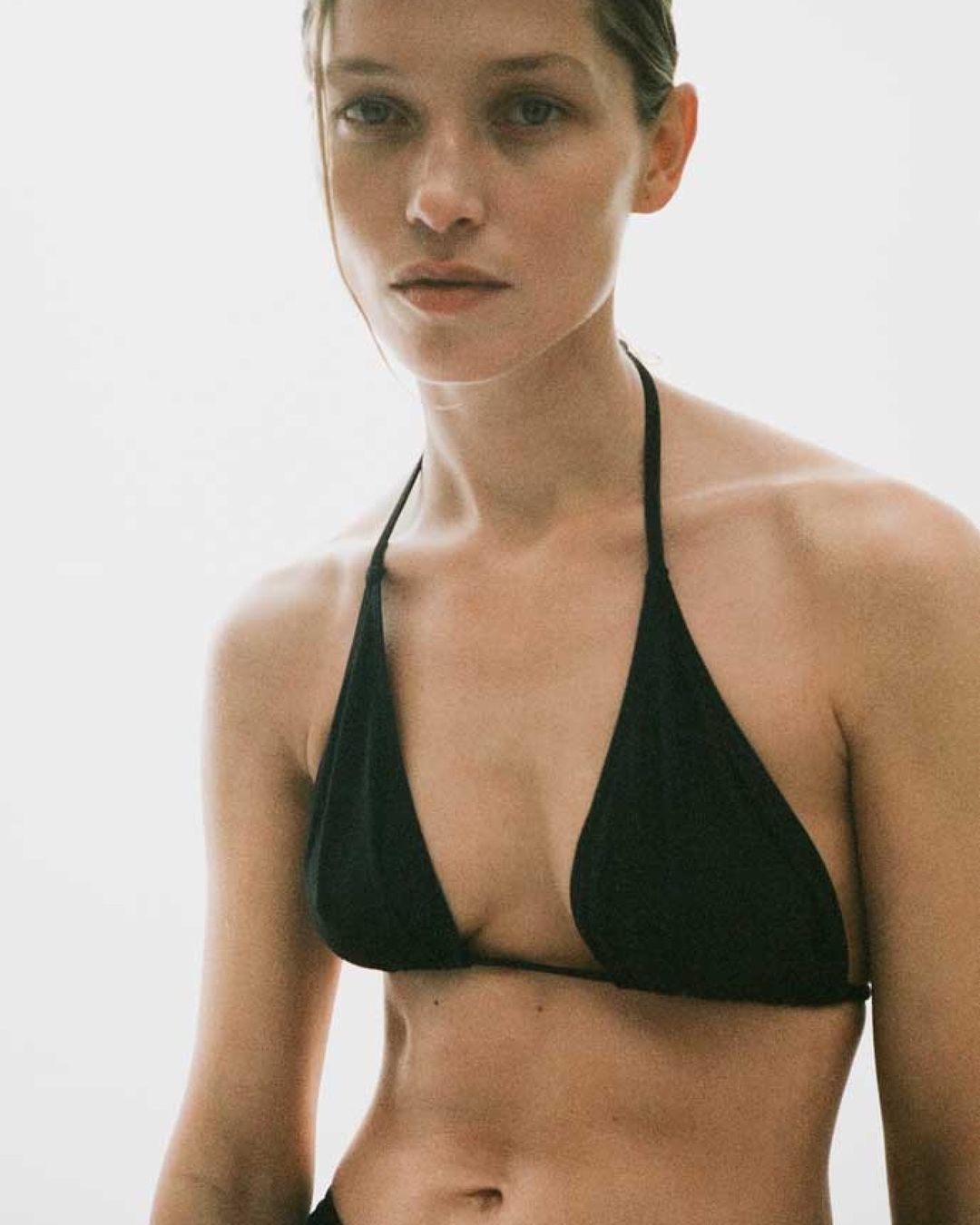 Close-up studio shot of a fair-skinned woman in a black bikini top against a bright white background, highlighting fashion and beauty.
