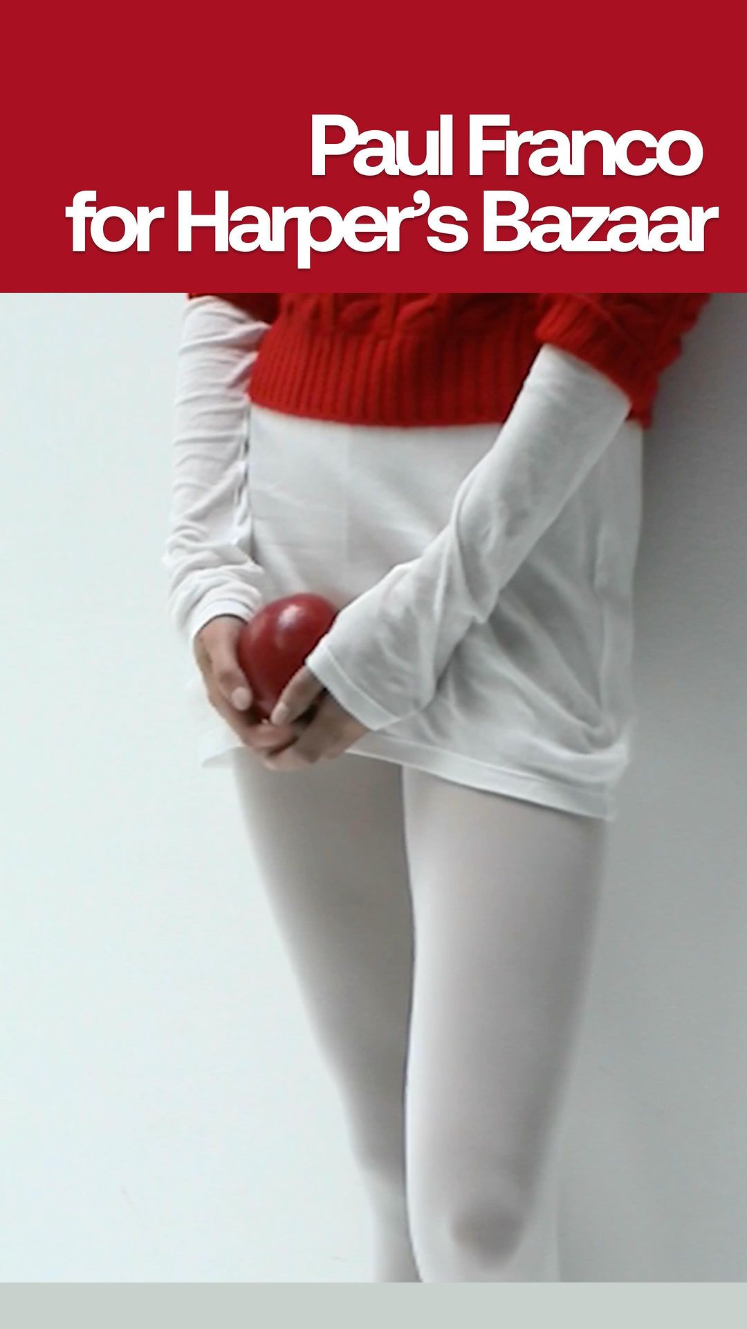Torso shot of a person in white tights holding a red apple, wearing a white long-sleeve and red short-sleeve top in a vertical portrait.
