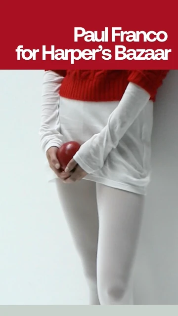 Torso shot of a person in white tights holding a red apple, wearing a white long-sleeve and red short-sleeve top in a vertical portrait.