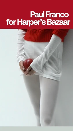 Torso shot of a person in white tights holding a red apple, wearing a white long-sleeve and red short-sleeve top in a vertical portrait.