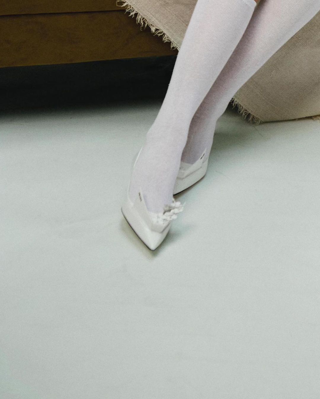 Close-up of a person wearing white knee socks and white pointed-toe heels indoors, set against a neutral-toned background.