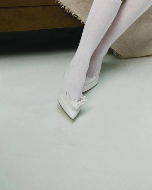 Close-up of a person wearing white knee socks and white pointed-toe heels indoors, set against a neutral-toned background.