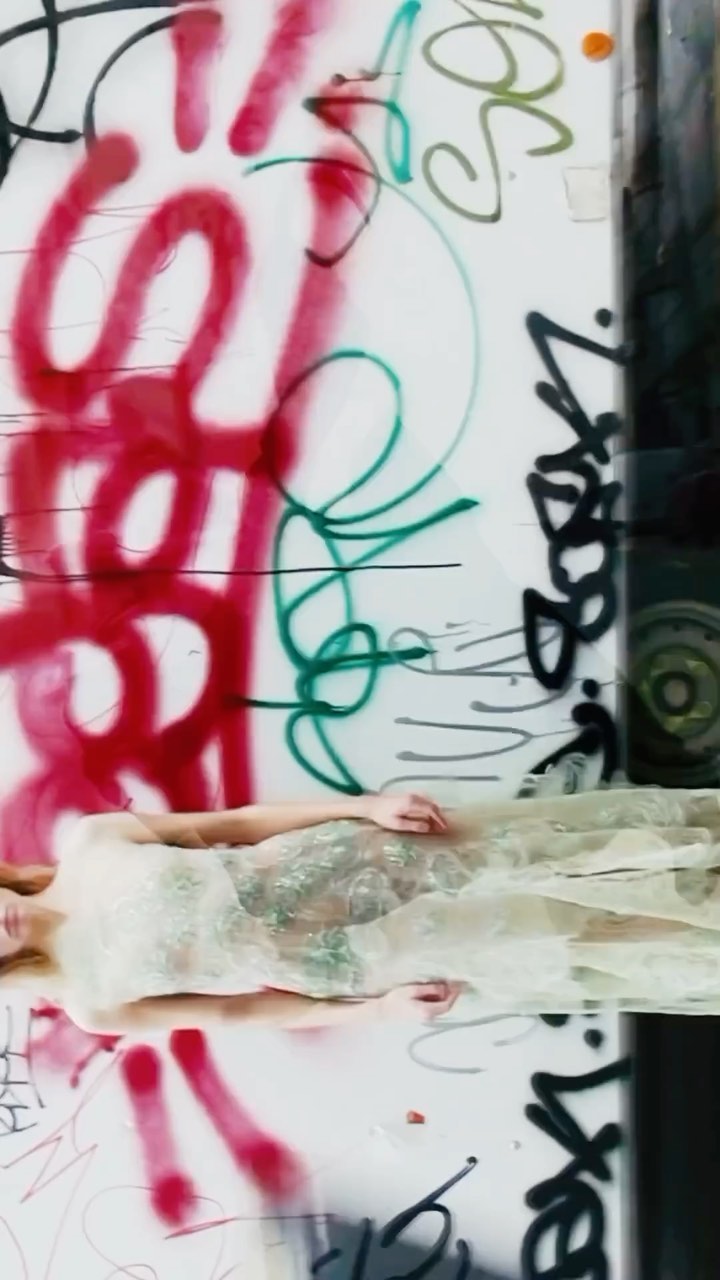 Woman in white dress laying in front of a graffiti wall with red, green and black spray paint in an indoor environment. 