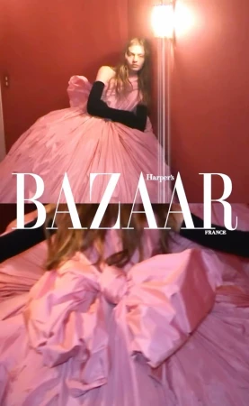 Woman in a voluminous pink gown and black gloves poses with a light fixture in a studio for Harper's Bazaar France fashion editorial.