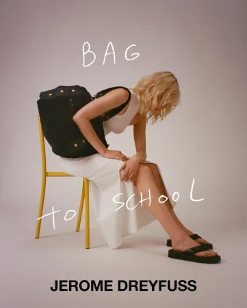 Blonde woman wearing white top and skirt seated on a yellow chair wearing black sandals, carrying a dark bag.