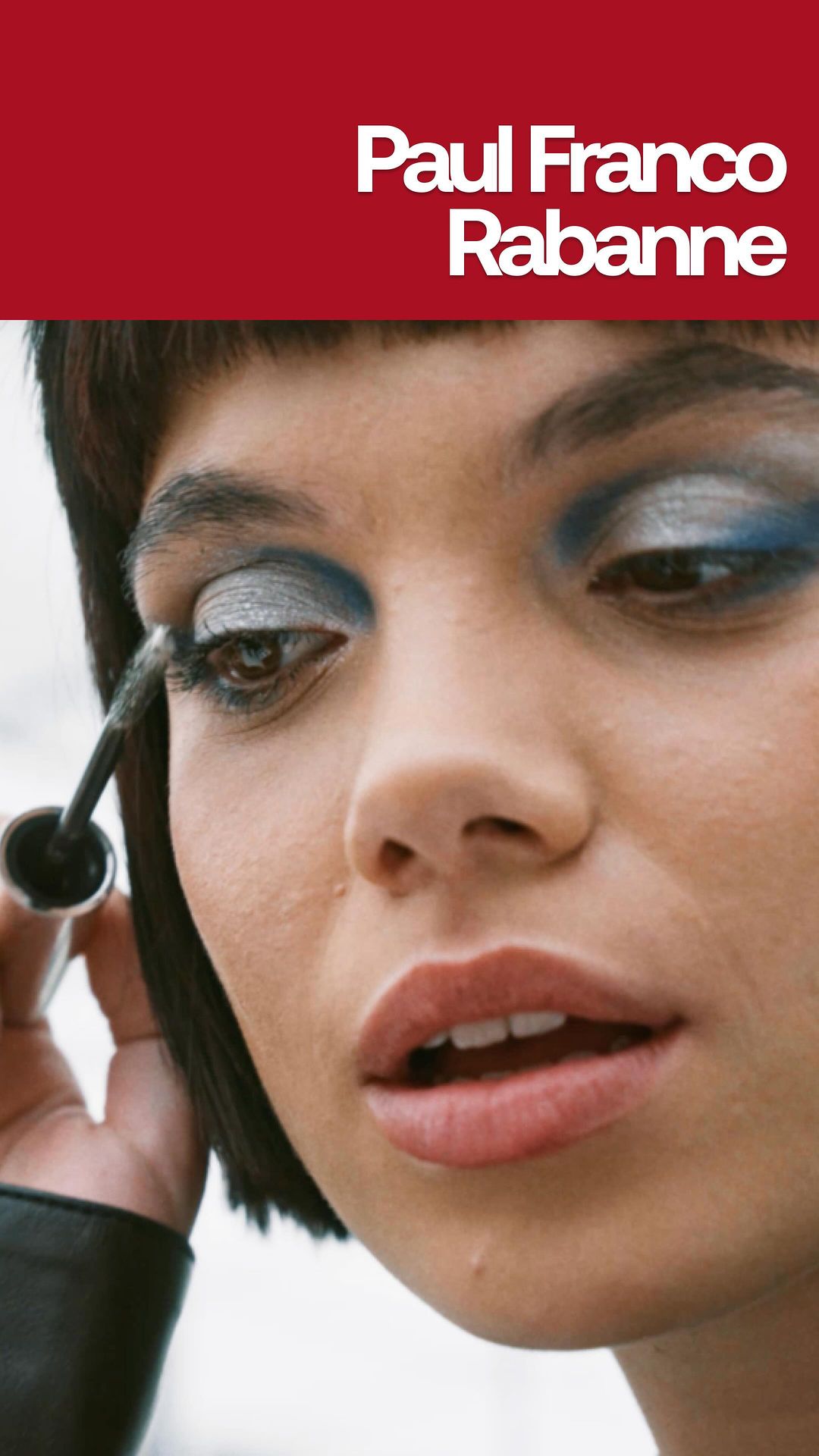 Close-up of a woman applying silver and blue eye makeup with mascara, against a blurred background and Paul Franco Rabanne text.