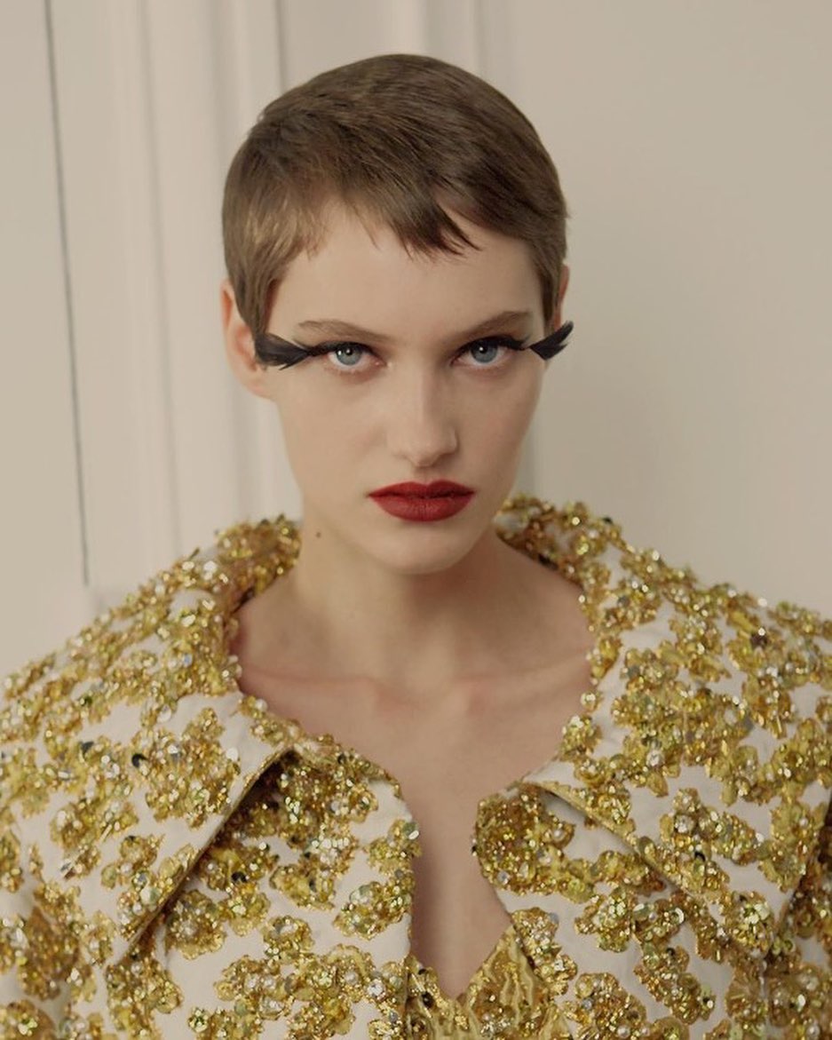 Close-up of a woman with fair skin and short brown hair wearing red lipstick and a jacket with gold embellishments looking at the camera.