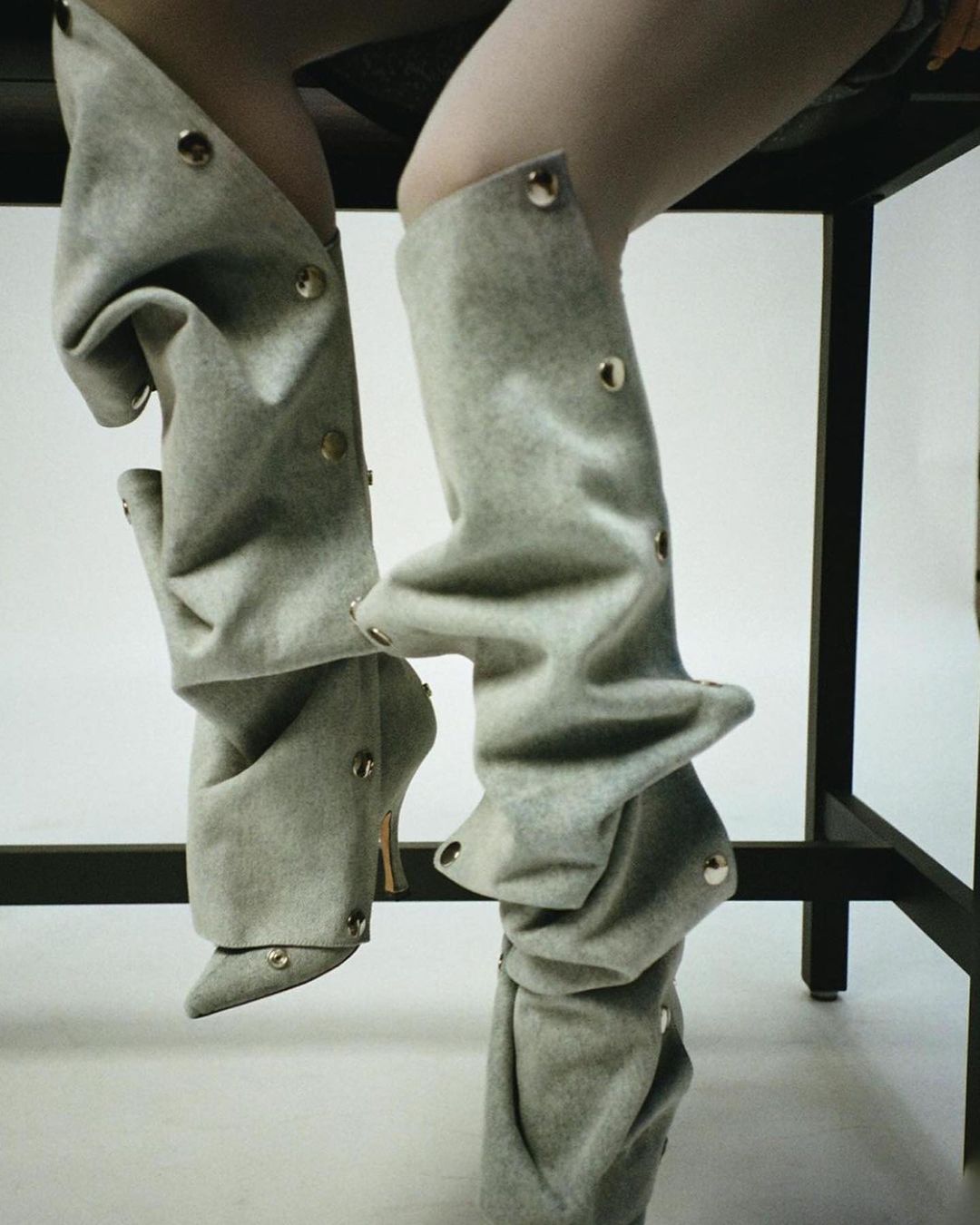 Close-up of a person wearing stylish, loose-fitting, gray leg coverings over high-heeled shoes, captured in a fashion-forward studio setting.
