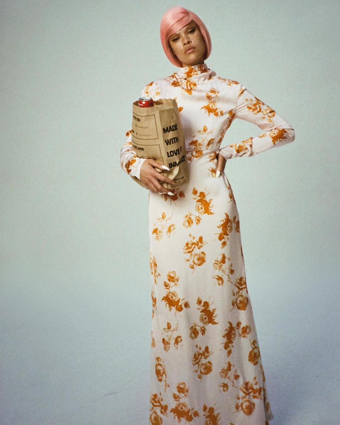 Full-length shot of a woman in a floral dress and pink wig holding a brown paper bag, posing against a pale blue background.