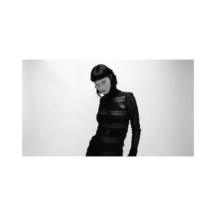 Black-and-white studio shot of a slender adult woman with short dark hair wearing a form-fitting, semi-sheer black top with cutout details.