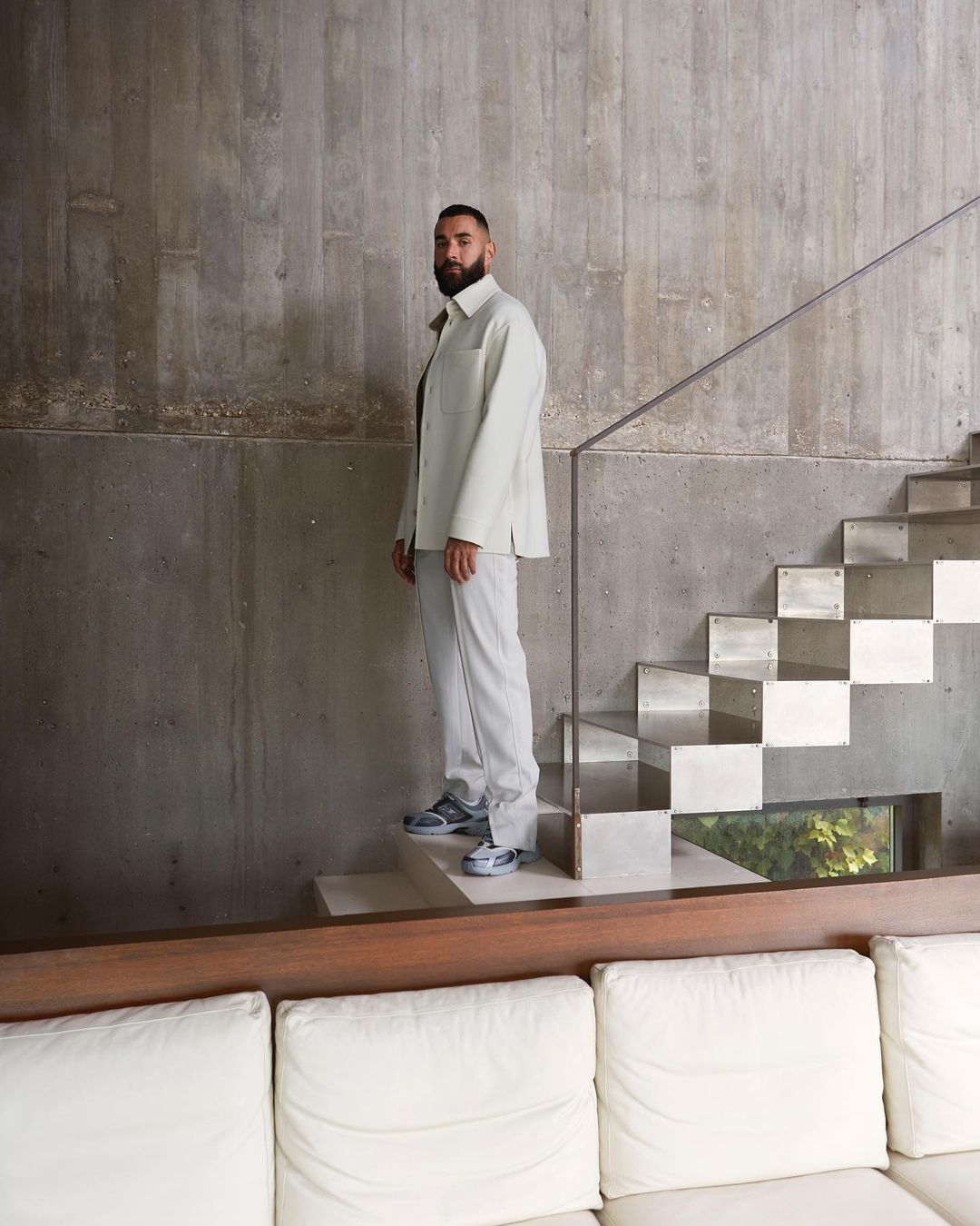 An adult male with a beard is standing on the steps of a staircase wearing a light colored suit, posing indoors in natural lighting.