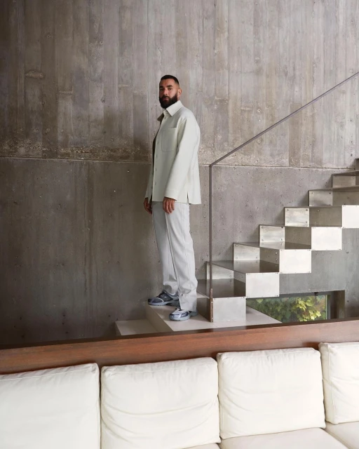 An adult male with a beard is standing on the steps of a staircase wearing a light colored suit, posing indoors in natural lighting.