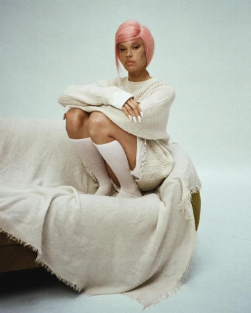 Woman with pink hair sits on a couch covered with a cream blanket in a white studio, showcasing fashion and beauty.