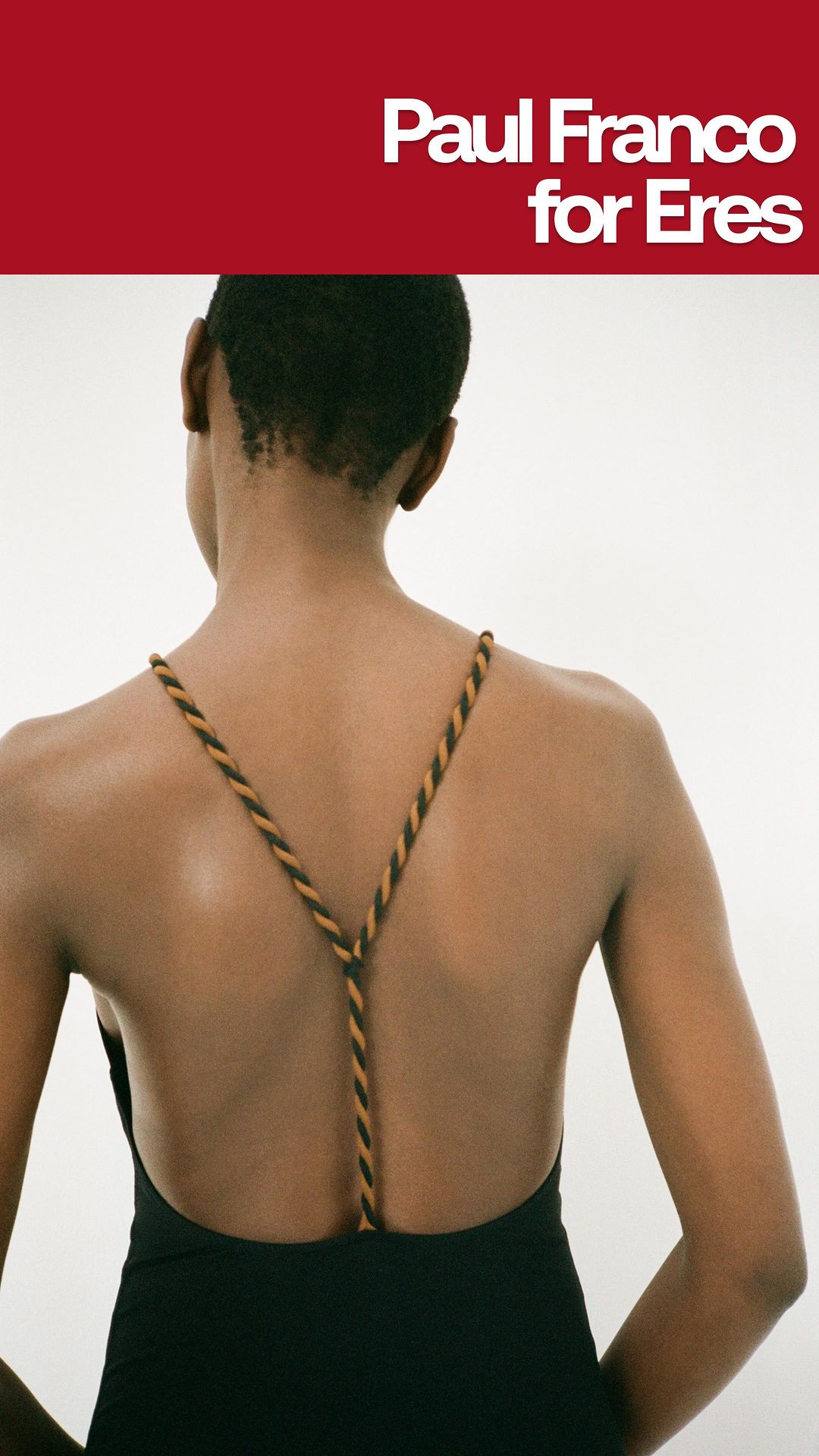 Back view of a Black woman wearing a black swimsuit with thin, beige and brown braided straps against a white background.