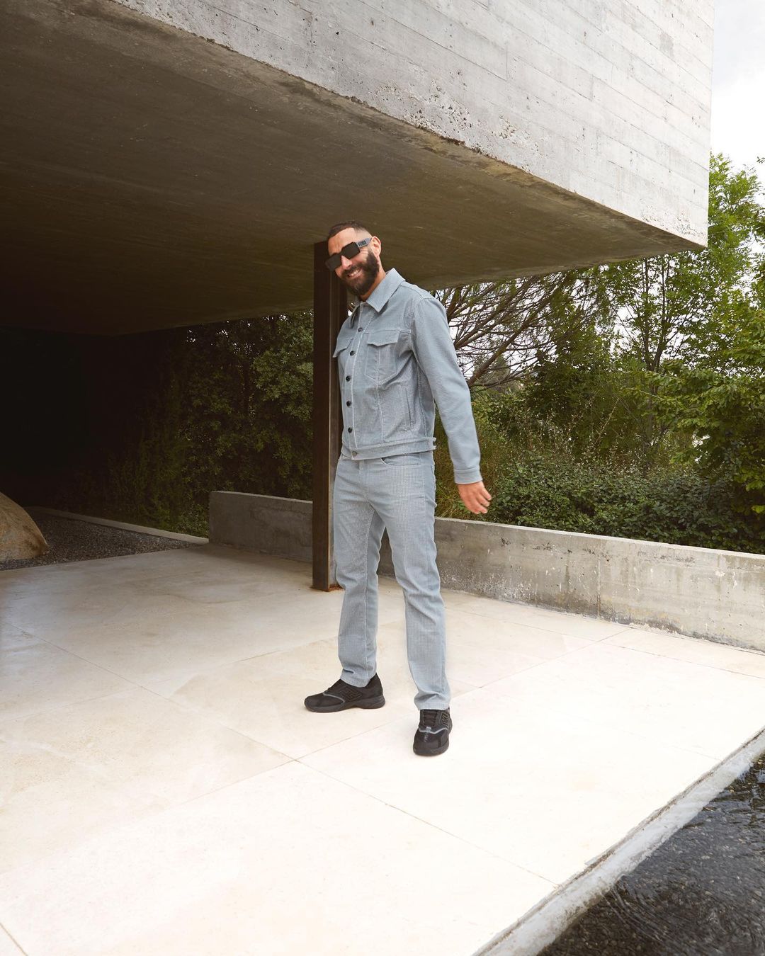 Bearded man in denim suit and sunglasses poses by concrete building in outdoor setting, showcasing a casual, modern style.