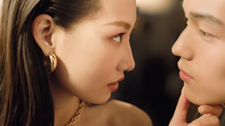 Close-up of an adult female and adult male gazing at each other, both wearing gold jewelry, against a warm, diffused background.