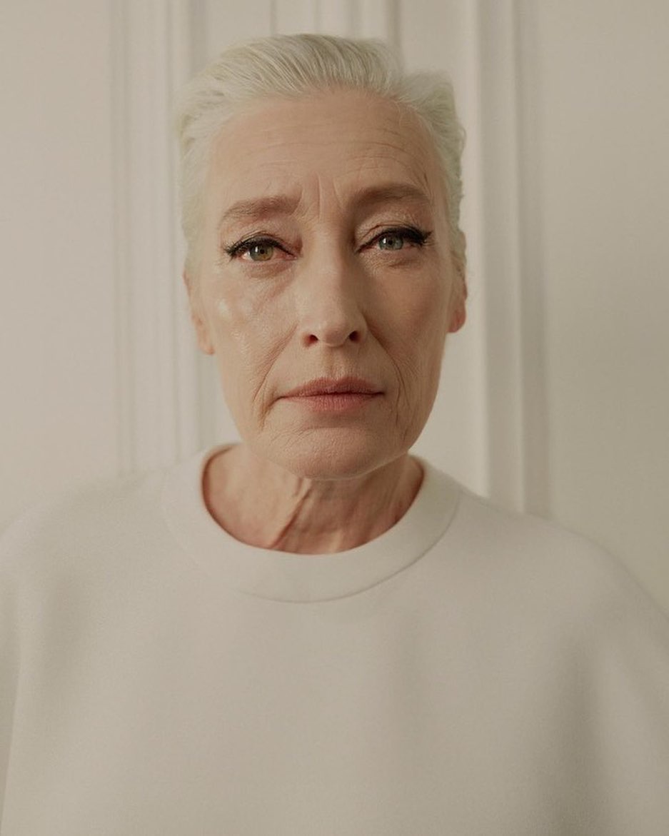 Close-up portrait of an adult woman with white hair wearing a white top against a bright neutral background indoors.
