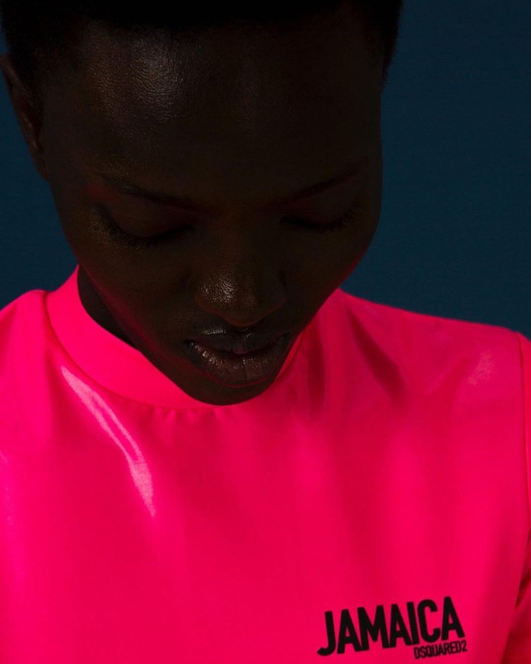 Close-up portrait of an adult woman with dark skin wearing a bright pink shirt with 'JAMAICA DSQUARED2' printed on the front against a blue background.