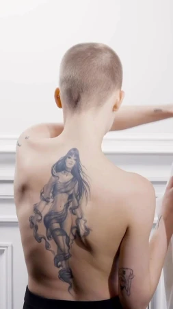 Back view of person with shaved head and a large tattoo of a woman on their back, posing indoors against a white paneled wall.