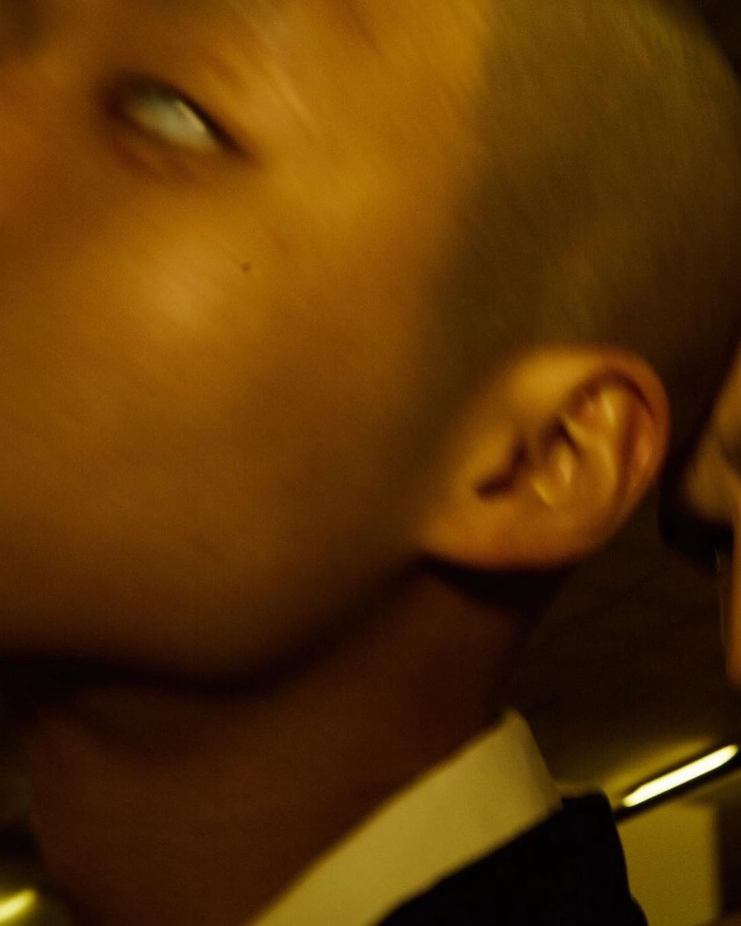 Close-up portrait of a short-haired person in profile wearing a dark jacket and white shirt under warm golden light by Paul Franco.
