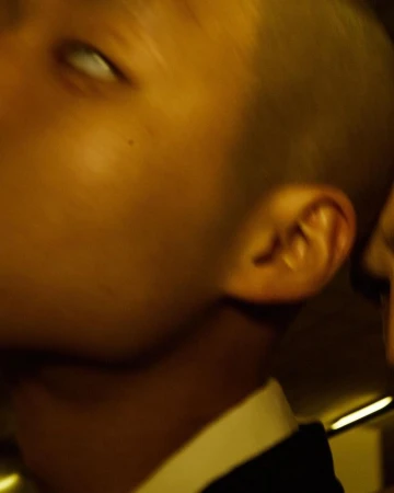 Close-up portrait of a short-haired person in profile wearing a dark jacket and white shirt under warm golden light by Paul Franco.